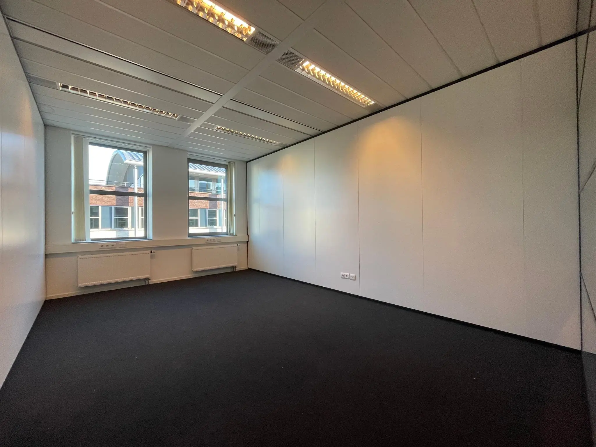 Lege kantoorruimte met witte wanden, zwarte vloerbedekking en twee ramen met uitzicht op een aangrenzend gebouw.